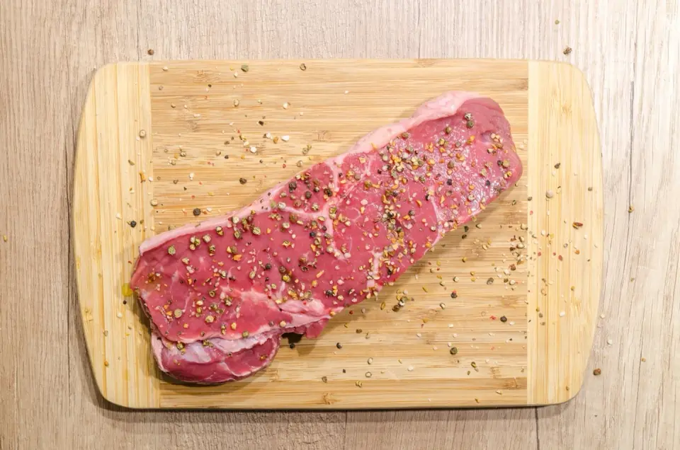 A raw beef steak seasoned with peppercorns on a pale chopping board